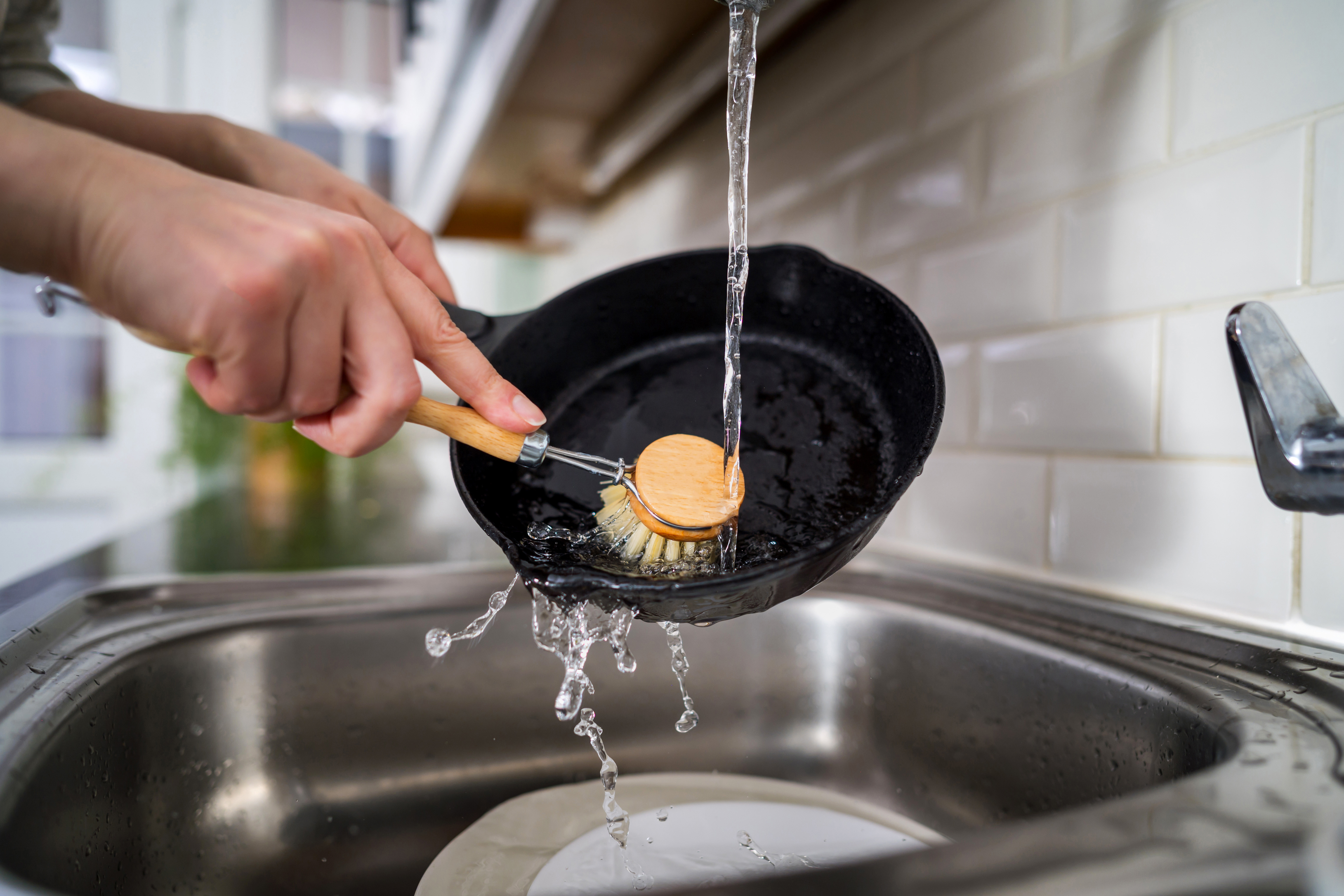 Scrubbing a cooking pan with a brush under running water to remove grease in a kitchen sink